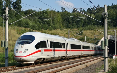 Fehér, nagysebességű ICE vonat halad zöldellő tájon, elektromos vezetékek alatt.