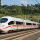 Fehér, nagysebességű ICE vonat halad zöldellő tájon, elektromos vezetékek alatt.